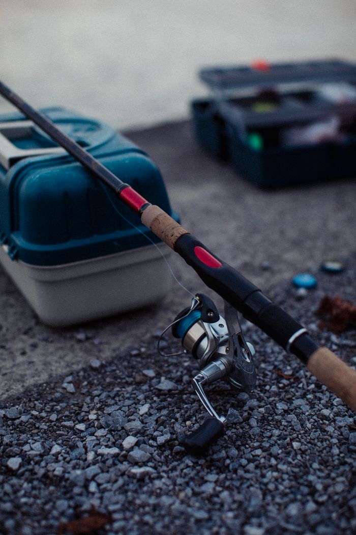 Close-up of a fishing rod and tackle box on a gravel surface, perfect for fishing enthusiasts.